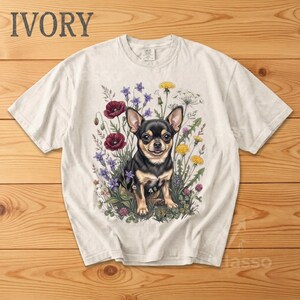 May include: Ivory-colored t-shirt featuring a detailed illustration of a Chihuahua dog amidst colorful wildflowers. The dog has black and tan fur, with the word "IVORY" printed at the top. The shirt is on a wooden surface.