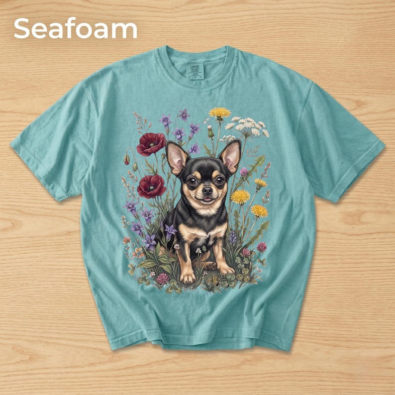 May include: Seafoam-colored t-shirt featuring a black and tan Chihuahua sitting amidst a colorful floral arrangement of red poppies, purple wildflowers, and yellow dandelions. The shirt has a relaxed fit and short sleeves, perfect for casual wear.