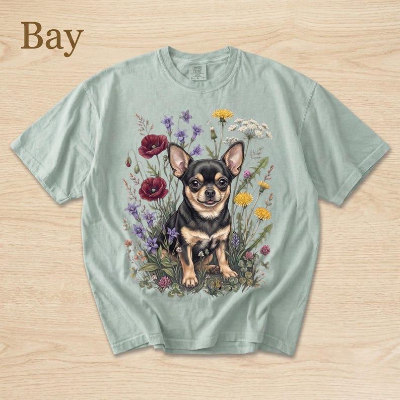 May include: A light green t-shirt featuring a detailed illustration of a black and tan Chihuahua sitting amidst wildflowers. The flowers include red poppies, yellow dandelions, and purple blooms. The word "Bay" is printed in the upper left corner.