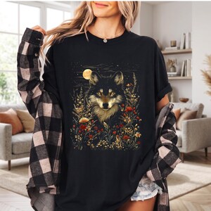 May include: Black t-shirt with a wolf design. The wolf has black fur and golden eyes, set against a backdrop of flowers and a full moon. The shirt is a relaxed, casual style.