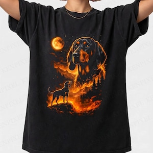 May include: Black t-shirt featuring a graphic of a dog with a fiery background, a full moon, and a smaller dog silhouette. The dog's head is detailed with black and brown fur. The design has orange and yellow hues.