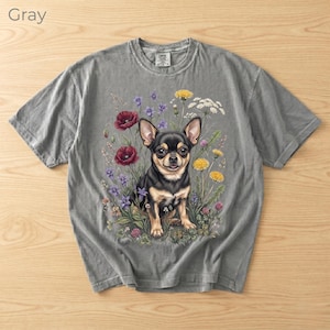 May include: Gray t-shirt featuring a detailed illustration of a Chihuahua dog surrounded by colorful wildflowers. The dog is black and tan, with large ears and a friendly expression. The word "Gray" is printed in the upper left corner.