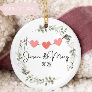 May include: A round white ceramic ornament featuring a printed wreath of green pine branches and red berries. Three pink hearts are strung on a line above the black cursive text Jason & Mary and the year 2026. A gold string is attached to the top.