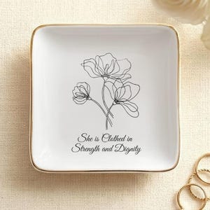 May include: A square white ceramic trinket dish with a gold-toned rim. The center features a black line art drawing of three flowers and the text "She is Clothed in Strength and Dignity" printed in a black script font on the smooth surface.