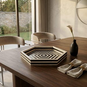 May include: A hexagonal tray featuring a repeating black and white geometric pattern on its surface and sides. The tray is placed on a wooden table alongside a small dark vase containing a single flower and two folded napkins with gold rings.