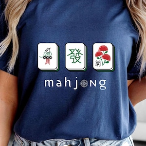 以下が含まれることがあります： マージャンのデザインが施されたネイビーブルーのTシャツ。グラフィックには、イラストと漢字が描かれた3つの牌と、白い文字で「mahjong」という単語が含まれています。このシャツは半袖のクルーネックです。