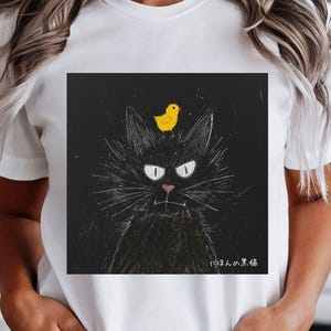 以下が含まれることがあります： 黒猫のイラストと黄色いヒヨコが頭に乗った白いTシャツ。猫は白い目とひげがあり、背景は黒です。「にほんの黒猫」の文字が猫の下に印刷されています。