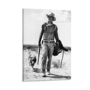 May include: Monochrome canvas print depicting a cowboy in a desert setting, carrying a rifle and saddle. A dog walks beside him. The cowboy is dressed in a hat, shirt, and jeans. The image is a vintage photograph.