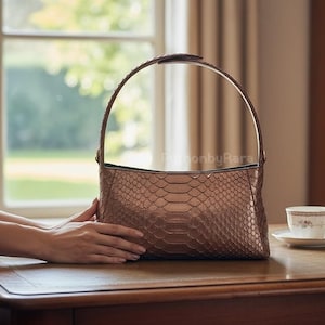 May include: A brown snakeskin-patterned handbag with a curved handle. It sits on a wooden table, alongside a teacup and saucer, and a notebook with a pen.