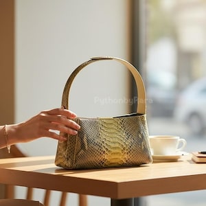 May include: A snakeskin-patterned handbag in green and yellow tones rests on a wooden table. The bag features a curved handle and a light-coloured interior. A coffee cup and notebook are also on the table.
