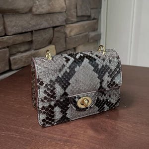 Może przedstawiać: Mała, prostokątna torebka ze wzorem skóry węża w odcieniach szarości, czerni i bieli. Torebka ma złote zapięcie i łańcuszkowe paski. Torebka leży na ciemnobrązowej drewnianej powierzchni.