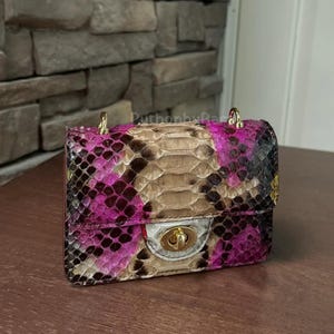 May include: A small, rectangular handbag with a snakeskin pattern in shades of pink, brown, and black. The bag has a gold-coloured chain strap and a clasp closure. The bag is on a wooden surface.