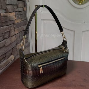 May include: A green and brown snakeskin-patterned shoulder bag with a black strap and gold-coloured hardware. The bag has a zipped main compartment and a front zipped pocket. The bag is on a wooden surface.