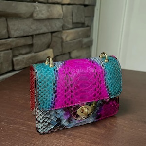 May include: A small, colourful handbag with a snakeskin pattern. The bag features a vibrant mix of colours, including pink, teal, and blue, with gold-toned hardware. The bag has a flap closure and a chain strap.