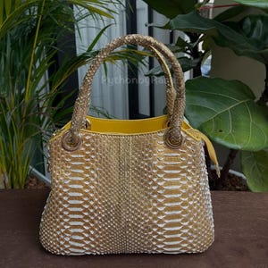 May include: A gold and white snakeskin handbag with a yellow interior. The bag has two braided handles and gold hardware. The bag is on a wooden surface with green plants in the background.