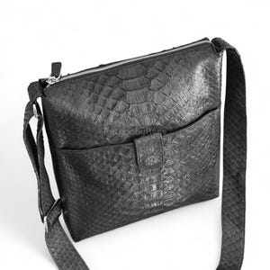 May include: Black leather crossbody bag with a textured, reptile-like pattern. The bag has a front pocket with a flap closure, a zippered top, and an adjustable shoulder strap. The bag is designed for everyday use.