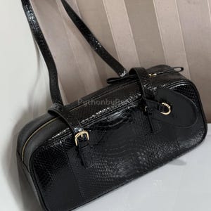 May include: A black rectangular handbag with a textured, snake-like pattern. The bag features a long shoulder strap, a top zipper, and gold-coloured hardware, including buckles and a circular clasp. The bag is approximately 30 cm long.