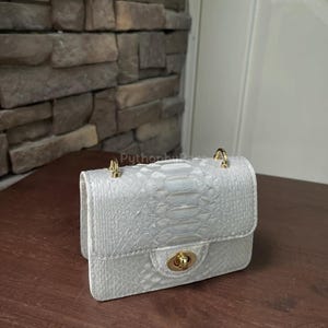 May include: A small, white handbag with a snakeskin pattern. The purse has a gold-coloured clasp and chain attachments. The bag is sitting on a wooden surface.