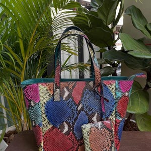 May include: A vibrant tote bag and purse set with a snakeskin design. The bag displays a mix of colours, such as red, blue, pink, and yellow. The bag has two handles and a zip closure. The purse also has a zip closure.