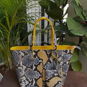 Może przedstawiać: Torba typu tote i pasująca do niej kosmetyczka z wzorem skóry węża w odcieniach szarości, czerni i beżu. Torba ma żółte wykończenia i uchwyty. Torba stoi na drewnianej powierzchni.