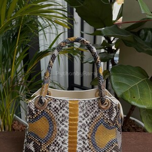May include: A snakeskin-patterned handbag with a cream interior. The bag features a mix of grey, gold, and blue scales, with a vertical gold stripe down the centre. The handles are also snakeskin, and the bag has gold-coloured hardware.