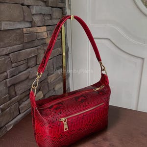 May include: A red snakeskin-effect shoulder bag with gold-tone hardware. The bag has two zipped compartments and a red velvet handle. The bag is displayed on a wooden surface.