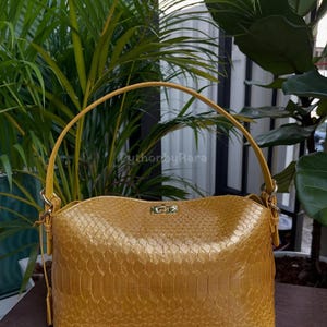 May include: Mustard yellow handbag with a snakeskin texture. The bag features a curved top, a structured rectangular base, and a single handle. Gold-coloured hardware accents the bag, including the handle attachments and a small clasp. The text "authorbyRara" is visible.