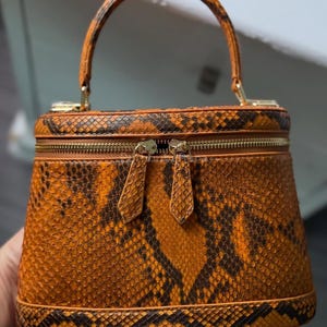 Könnte beinhalten: Eine kleine, orangefarbene und braune Handtasche mit Schlangenmuster, runder Form und einem oberen Griff. Die Tasche verfügt über einen goldenen Reißverschluss und goldfarbene Beschläge. Der Griff ist gebogen und mit goldenen Beschlägen an der Tasche befestigt.