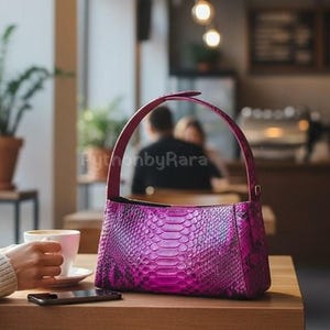 May include: A bright magenta handbag with a snakeskin pattern is on a wooden table. The bag has a curved handle and a rectangular shape. A person's hand holds a cup of coffee next to the bag. The text "PythonbyRara" is visible.
