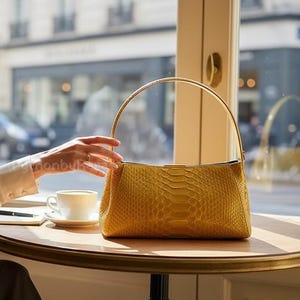 May include: A mustard yellow handbag with a snakeskin texture sits on a round table. The bag has a gold handle and a rectangular shape. A white teacup and saucer are on the table.