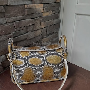 May include: A rectangular handbag with a snakeskin pattern in gold, grey, and white. The bag has a white strap and gold-coloured handles with gold rings. The bag is on a wooden surface.