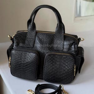 May include: Black leather handbag with a textured snakeskin pattern. The bag has two front pockets with gold zippers, a top handle, and a detachable shoulder strap. The hardware is gold-toned.