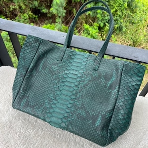 Peut inclure: Un grand sac fourre-tout vert émeraude avec un motif peau de serpent. Le sac a deux poignées et une forme rectangulaire. Le sac est fait d'un matériau texturé et semble être posé sur une surface blanche.
