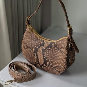 May include: A brown and tan snakeskin-patterned handbag with a gold zip and hardware. The bag has a curved shape with a short handle and a detachable shoulder strap. The bag is on a white surface.