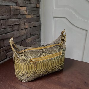 Puede incluir: Un bolso de hombro con estampado de piel de serpiente en tonos verdes, marrones y dorados. El bolso tiene forma rectangular con la parte superior curvada y cremalleras y herrajes dorados. Está sobre una superficie de madera.