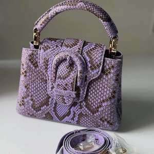 May include: A small, structured handbag in a lavender and brown snakeskin pattern. The bag features a top handle, a flap closure with a buckle, and a detachable shoulder strap. Gold-toned hardware accents the bag.