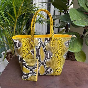 Op de afbeelding: Een gele tote bag en bijpassende portemonnee met een slangenprint. De tas heeft een gestructureerde vorm met een gele binnenkant en handvatten. De portemonnee is rechthoekig met een ritssluiting. De set wordt gepresenteerd op een houten oppervlak.