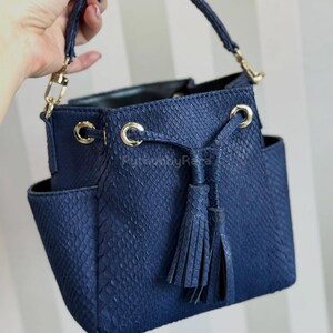 Może przedstawiać: Granatowa torebka z fakturą skóry węża. Torba ma górny uchwyt, złote okucia i detal z frędzlami. Posiada dwie zewnętrzne kieszenie i zapięcie na sznurek.