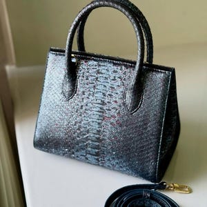May include: A small, structured handbag with a textured, iridescent finish in shades of black, blue, and hints of red. It features two rounded top handles and a detachable shoulder strap with gold-tone hardware. The bag is designed with a rectangular shape.