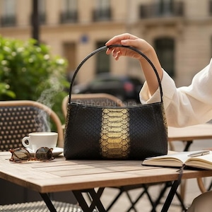 May include: Black handbag with a gold and black snakeskin pattern. The bag has a curved handle. A cup of coffee, sunglasses, and a notebook are on a wooden table. The scene is outdoors.