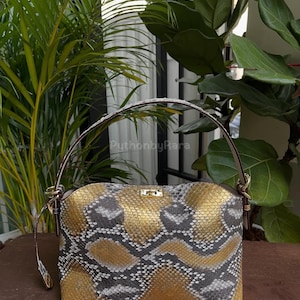 May include: A rectangular handbag with a gold, gray, and white snakeskin pattern. The bag has a brown handle and gold-colored hardware. The bag is sitting on a wooden surface, with green plants in the background.