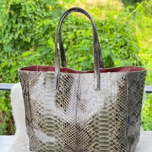 Puede incluir: Un bolso tote grande con un estampado de piel de serpiente en tonos grises, marrones y verdes. El bolso tiene dos asas y un interior rojo. El bolso está sobre una superficie blanca y esponjosa.