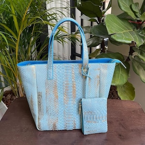 May include: A light blue tote bag and matching wallet set. The bags have a snakeskin pattern with gold accents. The tote has two handles and a zip closure. The wallet has a zip closure. The set is displayed on a wooden surface.