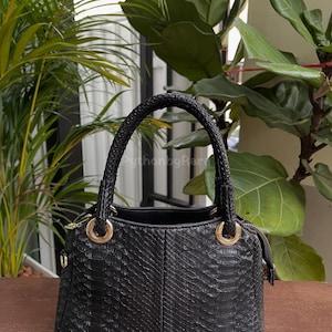 Puede incluir: Bolso de mano negro con un patrón texturizado similar a la piel de serpiente. El bolso tiene dos asas trenzadas y detalles metálicos dorados. El bolso está sobre una superficie de madera, con plantas verdes en el fondo.