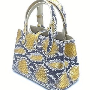 Puede incluir: Bolso estructurado con estampado de piel de serpiente en tonos dorados, grises y blancos. El bolso tiene dos asas superiores y un interior de color neutro. El diseño es de estilo tote clásico.