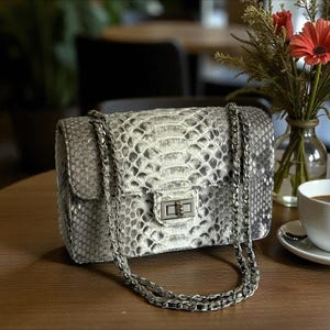 Könnte beinhalten: Rechteckige Handtasche mit weißem und grauem Schlangenmuster, silberner Kette und Verschluss. Im Hintergrund eine Vase mit Blumen und eine Tasse Kaffee auf einem Holztisch.