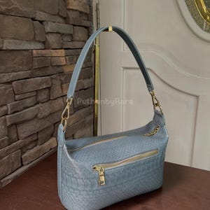May include: A light blue handbag with a textured, snake-skin pattern. The bag features a gold zip and hardware, and a matching shoulder strap. The bag is on a wooden surface.