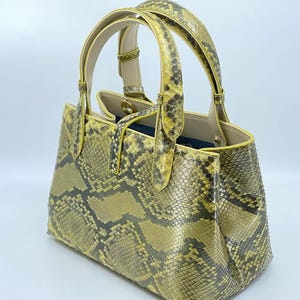 Puede incluir: Un bolso de mano amarillo y gris con estampado de piel de serpiente y dos asas. El bolso tiene forma rectangular con un diseño estructurado. Las asas y los ribetes son de color amarillo liso. El bolso está hecho de un material texturizado.