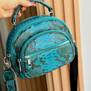 May include: A teal and black snakeskin-patterned handbag with silver zips and hardware. The bag has a top handle and a detachable shoulder strap. The bag is held by a hand.