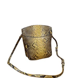 Può includere: Una piccola borsa a tracolla quadrata con un motivo a pelle di serpente nei toni del giallo, marrone e nero. La borsa ha una tracolla regolabile abbinata e una chiusura con fibbia. La borsa è realizzata in un materiale strutturato.